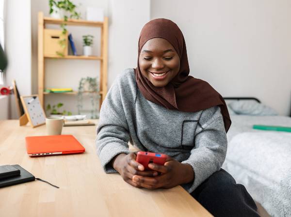 Smiling young woman of colour wearing headscarf or hijab browsing their smartphone while seated at desk in study room
